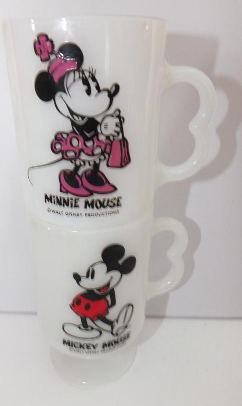 May include: Two white glass mugs featuring Minnie Mouse and Mickey Mouse. Minnie is depicted in a pink dress and bow, while Mickey wears red shorts. The mugs have heart-shaped handles and the text "Minnie Mouse" and "Mickey Mouse".