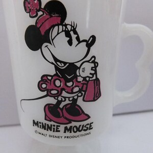 May include: White glass Minnie Mouse mug featuring a black and pink Minnie Mouse graphic. The mug has a heart-shaped handle and the words "Minnie Mouse" and "Walt Disney Productions" printed on the front.