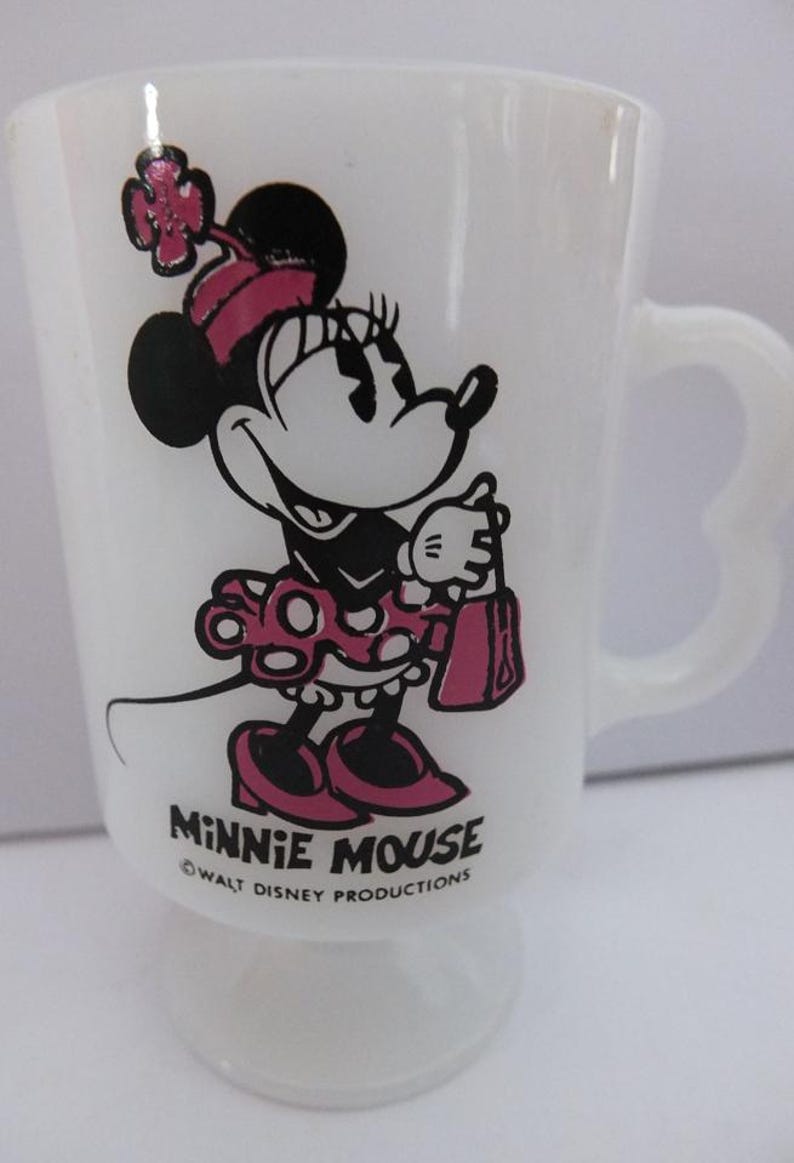 May include: White glass Minnie Mouse mug featuring a black and pink Minnie Mouse graphic. The mug has a heart-shaped handle and the words "Minnie Mouse" and "Walt Disney Productions" printed on the front.