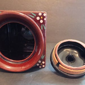 K&ouml;nnte beinhalten: Eine braune Keramikdose mit Deckel. Die Dose hat ein Blumenmuster mit wei&szlig;en Bl&uuml;ten und gr&uuml;nen Bl&auml;ttern. Der Deckel ist schwarz mit einem braunen Rand.