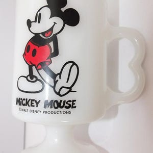 May include: White pedestal mug featuring Mickey Mouse. The cartoon character is depicted in black, red, and white. The mug has a curved handle and the words "Mickey Mouse" and "Walt Disney Productions" are printed on the front.