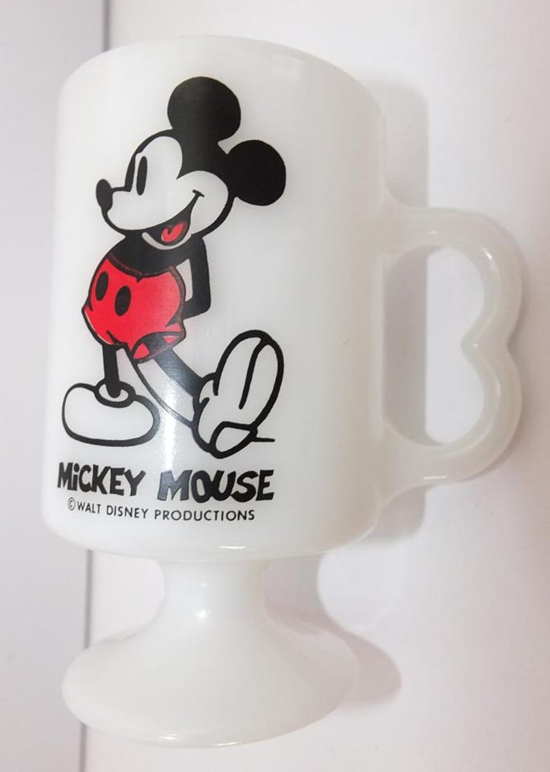 May include: White pedestal mug featuring Mickey Mouse. The cartoon character is depicted in black, red, and white. The mug has a curved handle and the words "Mickey Mouse" and "Walt Disney Productions" are printed on the front.