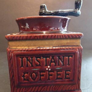 K&ouml;nnte beinhalten: Eine braune Kaffeem&uuml;hle aus Keramik mit einem schwarzen Griff. Die M&uuml;hle hat ein Etikett mit der Aufschrift "INSTANT COFFEE" mit gr&uuml;nen und wei&szlig;en Akzenten.