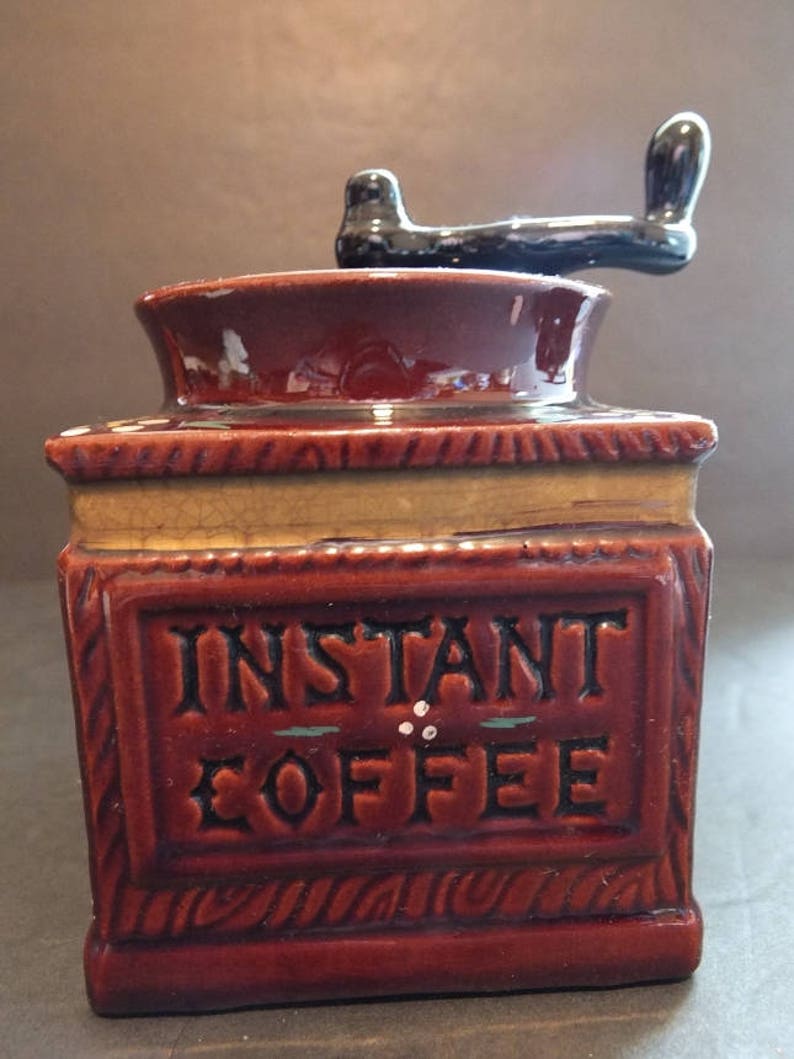 K&ouml;nnte beinhalten: Eine braune Kaffeem&uuml;hle aus Keramik mit einem schwarzen Griff. Die M&uuml;hle hat ein Etikett mit der Aufschrift "INSTANT COFFEE" mit gr&uuml;nen und wei&szlig;en Akzenten.
