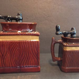 K&ouml;nnte beinhalten: Zwei Keramik-Kaffeem&uuml;hlen mit Holzmaserung. Die gr&ouml;&szlig;ere M&uuml;hle hat eine braune Basis und einen schwarzen Griff. Die kleinere M&uuml;hle hat eine braune Basis und einen schwarzen Griff. Beide M&uuml;hlen haben einen goldenen Rand.