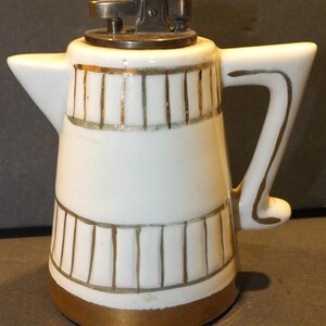 Könnte beinhalten: Weißer Keramik-Teekannen-förmiger Feuerzeug mit goldenen Verzierungen und einem Metalldeckel.