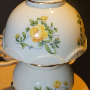 May include: A white porcelain table lamp featuring a rounded base and a matching dome-shaped shade. Both the base and the shade are decorated with a floral pattern of yellow roses and green leaves, accented by thin gold trim along the edges.