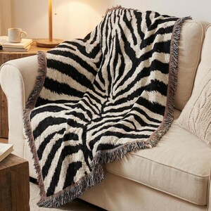 May include: A black and white zebra-striped throw blanket with fringed edges is draped over a beige sofa. The blanket's pattern is a bold, repeating design. The sofa has a neutral color palette, with a matching pillow.