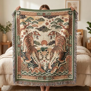 May include: A woven blanket with a detailed illustration of two tigers facing each other, with a mountain and sun in the centre. The blanket has a green border and fringe, with a beige background and brown, orange, and green accents.