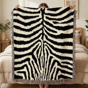 May include: A black and white zebra print throw blanket with fringed edges. The blanket features a bold pattern of black stripes on a cream-colored background, creating a striking visual contrast. This decorative blanket is suitable for a bed or sofa.
