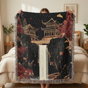 May include: A black throw blanket with a detailed illustration of an Asian landscape. The design features golden pagodas, a waterfall, and red and gold foliage. The blanket has a fringed edge.