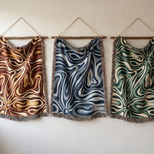 May include: Three decorative wall hangings with a swirling pattern in shades of brown, blue, and green. Each textile is fringed at the bottom and suspended from a wooden rod with a natural twine hanger. The abstract design adds a touch of modern art to any space.