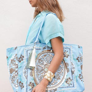 Peut inclure: Grand sac fourre-tout bleu clair avec un motif floral et géométrique détaillé dans les tons marron, blanc et bleu. Le sac a un pompon assorti et est porté par une personne portant une chemise bleu clair et un bracelet manchette doré.