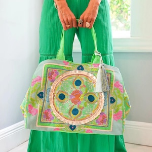 May include: A large, embroidered tote bag with a light green background and colorful floral and geometric patterns. The bag has green handles and a decorative tassel. The bag is held by a person wearing a green outfit.