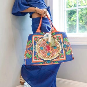 Peut inclure: Un grand sac fourre-tout bleu avec une bordure orange et un motif circulaire coloré. Le sac est orné d'un pompon décoratif et est porté par une personne vêtue d'une tenue bleue. Le motif du sac comprend des motifs floraux et géométriques.