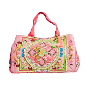 Peut inclure: Un sac fourre-tout rose avec un motif floral brodé. Le sac présente un motif circulaire central avec des accents roses, verts et jaunes, entouré d'une bordure florale rectangulaire. Il a des anses corail et un pompon.