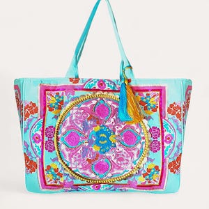 Peut inclure: Un sac fourre-tout turquoise avec un motif floral et géométrique coloré. Le sac présente un médaillon central avec des accents roses, bleus et dorés, et une pampille. Les poignées et la garniture sont turquoise.