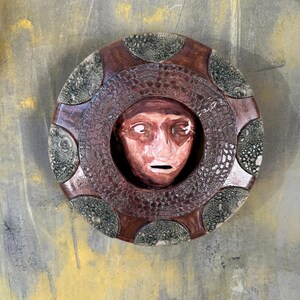 May include: A ceramic wall hanging featuring a sculpted face within a circular frame. The face is painted in shades of red and brown, with a neutral expression. The frame is a dark brown with a textured, lace-like pattern and green accents.