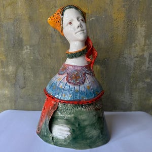 Può includere: Scultura in ceramica di una donna con volto bianco, occhi rossi e foulard arancione con motivi a stelle nere. La scultura ha un top blu e rosso fantasia e un vestito verde. È visibile una mano bianca.