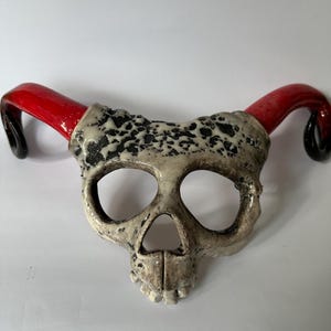 Op de afbeelding: Een keramisch schedelmasker met rode hoorns. De schedel is gebroken wit met zwarte gespikkelde details. De hoorns zijn glanzend rood, overgaand in zwart aan de uiteinden. Het masker heeft grote oogkassen en een gedetailleerde kaaklijn.