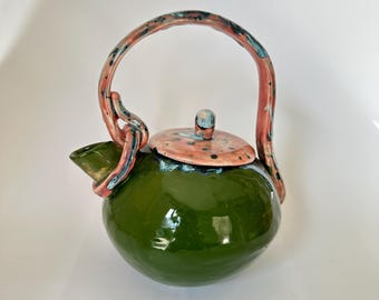 May include: A handcrafted ceramic teapot with a glossy green body, a speckled pink lid, and a curved handle. The handle and spout are accented with blue and pink hues. The teapot is a unique piece of kitchenware.