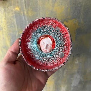 Op de afbeelding: Een handgemaakte keramische schaal met een centrale gezichtssculptuur. De schaal heeft een rood en turquoise kleurenschema met een textuur. Het gezicht heeft rood haar en een wit gezicht. De schaal wordt in een hand gehouden.