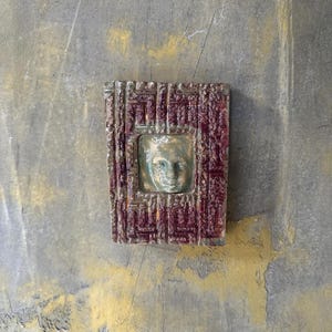 May include: A rectangular wall art piece featuring a face relief at its centre. The face is framed within a square, surrounded by a burgundy and gold patterned border. The artwork is mounted on a grey and yellow textured wall.