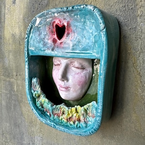 Könnte beinhalten: Keramik-Wandskulptur mit einem ruhigen Gesicht in einem türkisfarbenen Rahmen. Oben ist eine herzförmige Öffnung zu sehen, unten ein strukturierter, farbenfroher Rand. Die Skulptur ist an einer strukturierten, neutralfarbenen Wand befestigt.