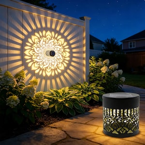 May include: A black solar-powered outdoor lantern with a decorative cut-out design, casting a warm light. A matching solar light is mounted on a white fence, projecting a floral pattern. The scene is set at dusk, surrounded by greenery and a stone pathway.