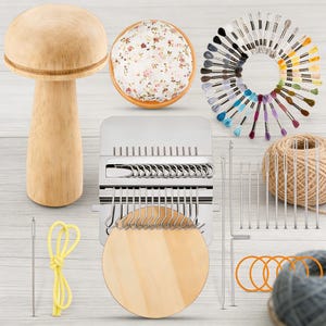 Könnte beinhalten: Eine Sammlung von Bastelmaterialien, darunter ein pilzförmiger Nadelhalter aus Holz, ein Blumen-Nadelkissen, ein Metallwebrahmen, Stickgarn, Nadeln und Wolle. Die Artikel sind auf einer hellen Oberfläche angeordnet.