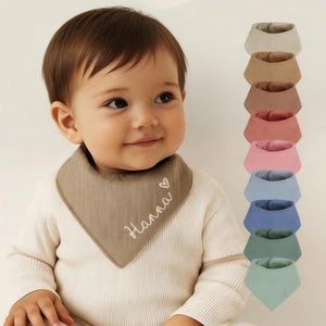 May include: A baby wearing a taupe bandana bib with the name "Hanna" embroidered in white. A stack of seven bibs in various colors, including beige, brown, pink, blue, and green, is displayed to the right. The bibs are made of soft fabric.