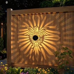 May include: A wooden fence with a solar-powered light fixture. The light projects an eagle design with radiating beams. The fixture is circular and black, mounted in the center of the eagle design. The fence is made of vertical wooden planks.