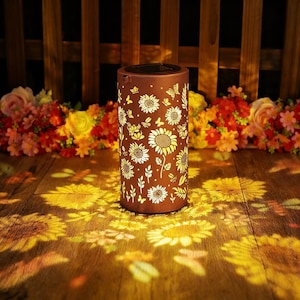Puede incluir: Una linterna solar cilíndrica marrón con un diseño de girasoles y mariposas. La linterna proyecta un diseño de girasol sobre la superficie de madera que hay debajo. El fondo presenta una valla de madera y flores coloridas.