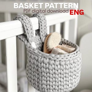 Könnte beinhalten: Ein handgehäkelter, grauer Korb hängt an einer weißen Gitterbett-Reling. Im Korb befinden sich eine Holzbürste und gefaltete Babyartikel. Der Text "BASKET PATTERN PDF digital download ENG" ist oben im Bild zu sehen.