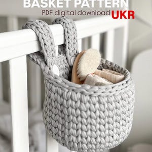 May include: A gray, crocheted basket hanging from a white crib rail. The basket contains a wooden brush and folded cloths. The text "BASKET PATTERN PDF digital download UKR" is at the top of the image.