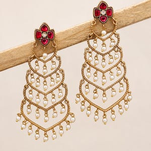 Peut inclure: Une paire de boucles d'oreilles chandelier dorées avec une structure en gradins en forme de chevron. Chaque boucle est ornée de petites perles blanches suspendues aux bords et d'un motif floral supérieur serti de quatre pierres rouge foncé.