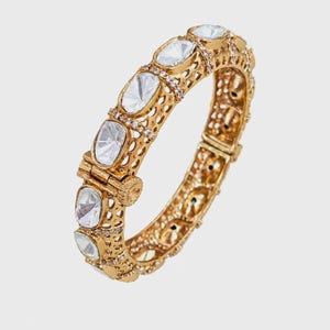 May include: A gold-toned hinged bangle bracelet featuring a series of large, oval-shaped clear stones set within an intricate, openwork metal lattice frame. The edges of the bracelet are accented with small, sparkling clear crystals.