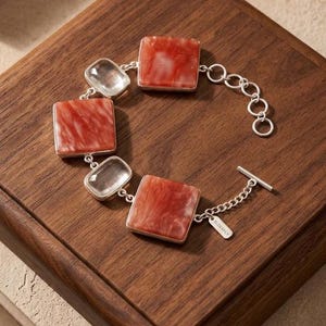 Pode incluir: Uma pulseira prateada com três elos quadrados de resina marmorizada laranja e branca, alternados com dois elos retangulares de vidro transparente. A peça inclui um fecho de barra e uma pequena etiqueta gravada com a palavra ELIDANCE.