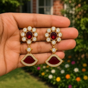 May include: A pair of gold-toned dangle earrings featuring a central oval deep red stone surrounded by a floral arrangement of small clear round crystals, with a fan-shaped pendant at the base containing a matching red stone and crystal border.