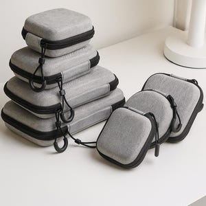 以下が含まれることがあります： 黒いジッパーとコードが付いた、さまざまなサイズのグレーの収納ケースのコレクション。ケースは長方形と正方形で、白い表面に積み重ねて配置されています。