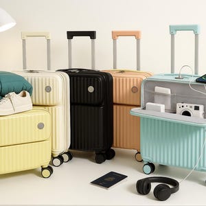 Può includere: Una collezione di valigie con ruote colorate in varie tonalità, tra cui giallo, nero, pesca e verde acqua. Una valigia è aperta, rivelando una fotocamera e un telefono. Una coperta e delle scarpe poggiano su una valigia gialla. Sono visibili anche un passaporto e delle cuffie.