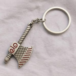 May include: Silver-colored axe keychain with a detailed design. The axe head features red accents and a crosshatch pattern. The handle has a textured, twisted design. Includes a silver key ring.