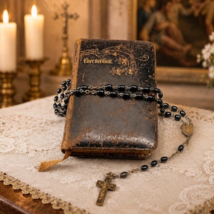 Könnte beinhalten: Ein antikes, abgenutztes Buch mit schwarzem Ledereinband und der goldenen Prägung 'Ehre sei Gott' auf dem Deckel, umwickelt mit einem schwarzen Perlenrosenkranz mit Metallkruzifix. Das Buch hat eine gealterte Textur und ein kleines Stofflesezeichen.