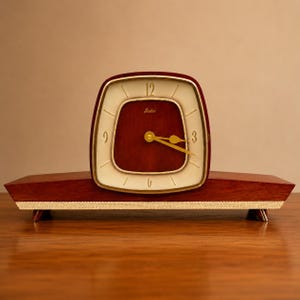 May include: A mantel clock with a dark wood base and a square-shaped face featuring a cream-colored dial, gold-toned hands, and the brand name Lasko printed in gold. The clock has a textured gold-colored trim along the bottom of the base.