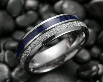 Könnte beinhalten: Nahaufnahme eines silbernen Rings mit blauem Steineinsatz und einem strukturierten, silbernen Band. Der Ring ist vor einem dunklen Hintergrund aus glatten, schwarzen Steinen platziert. Das Design des Rings weist eine polierte Oberfläche auf.