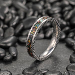 May include: A silver ring with a black inlay filled with colorful, iridescent fragments. The ring band has a textured, chevron-like pattern. The ring is displayed on a bed of dark, smooth stones.