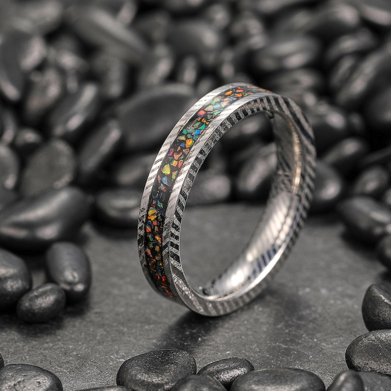 May include: A silver ring with a black inlay filled with colorful, iridescent fragments. The ring band has a textured, chevron-like pattern. The ring is displayed on a bed of dark, smooth stones.
