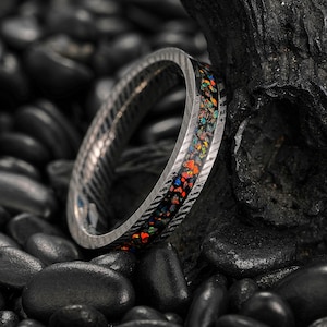 May include: A silver ring with a patterned band and a colorful inlay of orange, blue, and yellow stones. The ring is set against a backdrop of black stones, highlighting its intricate design and vibrant colors.