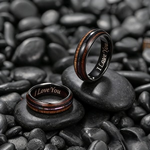 May include: Two black rings with the inscription "I Love You" are displayed on black stones. One ring features a wood grain design, while the other has a wood and purple inlay. The rings are likely wedding bands or promise rings.