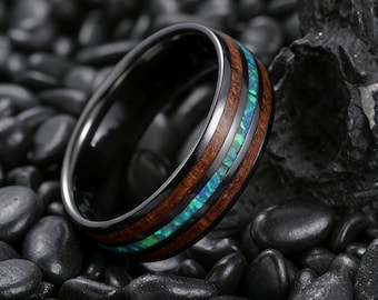 May include: A close-up of a black ring with a wood grain band and a vibrant blue-green opal inlay. The ring is set against a dark background of black stones, highlighting its polished finish and unique design.